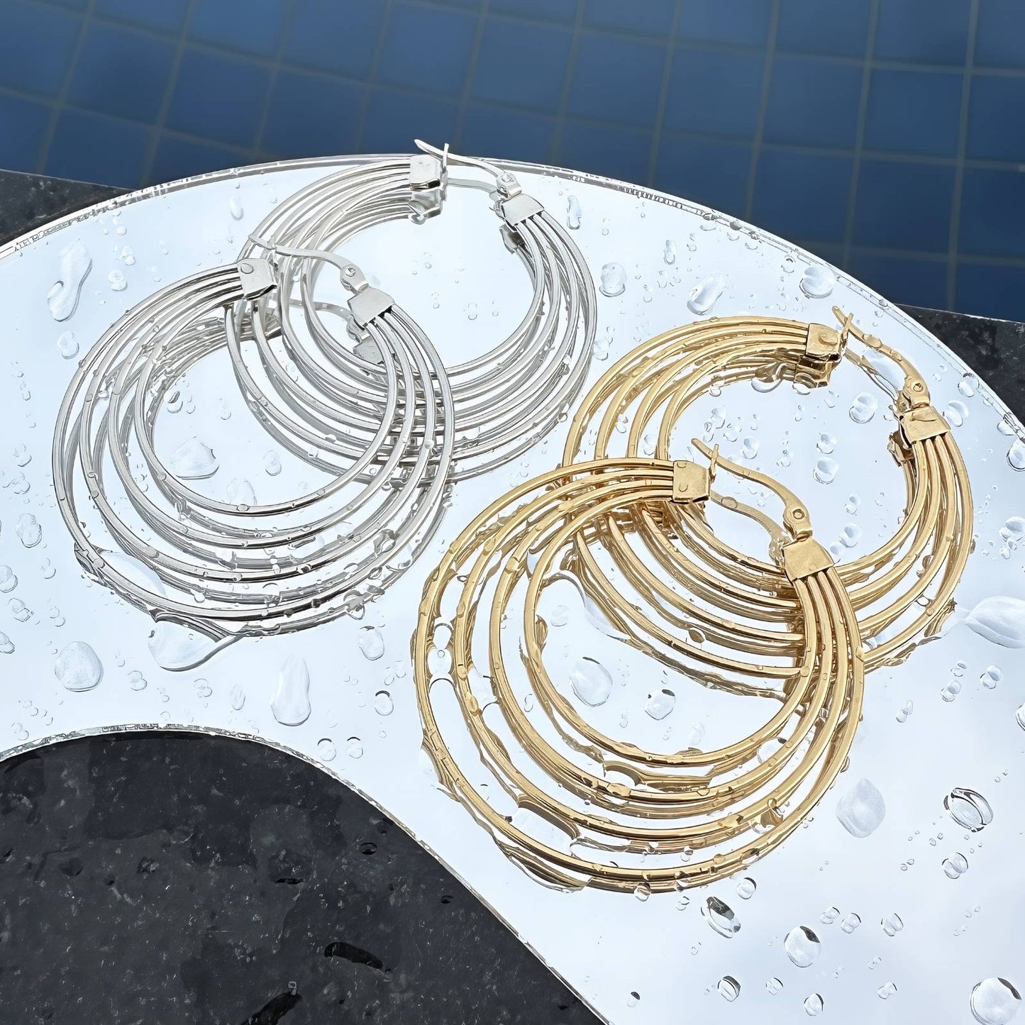 Pair of gold and silver hoop earrings on a wet surface with a blue tiled background.